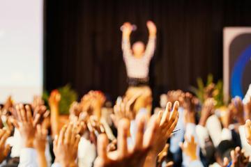 Blurry picture of audience raising hands in front of a stage where a speker stands. The photos is taken from the perspective of the audience.