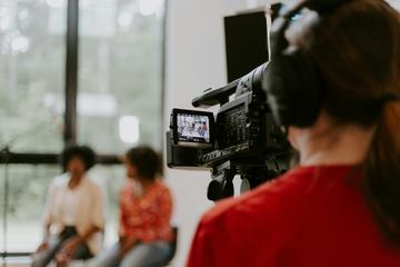 Man in red shirt holding black videocamera shooting two women in conversation.