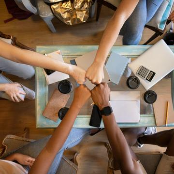 Four people bringing their fists together over a table with notepads and takeaway coffee cups