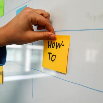 A hand sticking a note on a whiteboard with the words 'how to' written on it.