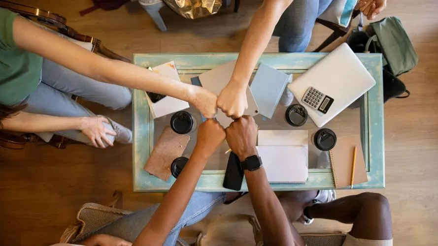 Four people bringing their fists together over a table with notepads and takeaway coffee cups