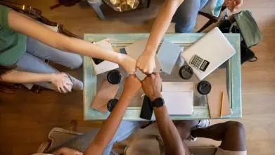 Four people bringing their fists together over a table with notepads and takeaway coffee cups
